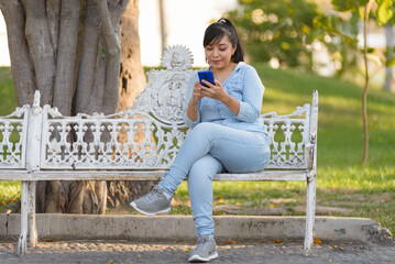 A woman sits on a white park bench, engrossed in her smartphone at sunset