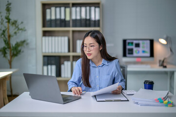A woman is sitting at a desk with a laptop and a stack of papers