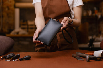 Tailor holds finished product black leather bag against background of his workplace in workshop