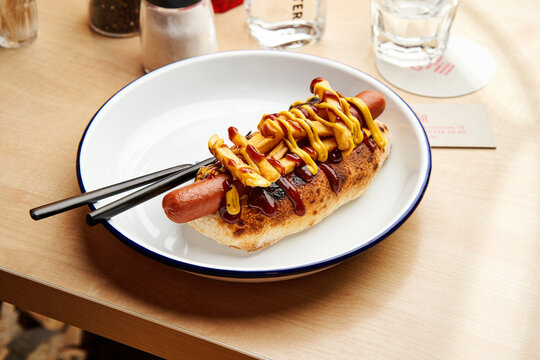 A hot dog garnished with mustard and ketchup is placed on a white plate with chopsticks beside it. The setting suggests a relaxed dining atmosphere with minimalist decor