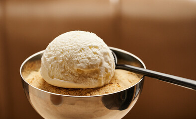 A generous scoop of vanilla ice cream sits atop a bed of biscuit crumbs in a silver bowl, accompanied by a sleek black spoon, creating a delightful dessert moment in a cafe
