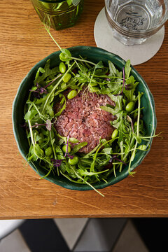 A delicious serving of beef tartare is placed in the center of a vibrant green salad composed of various fresh greens and herbs. The rustic bowl enhances the dish's appeal