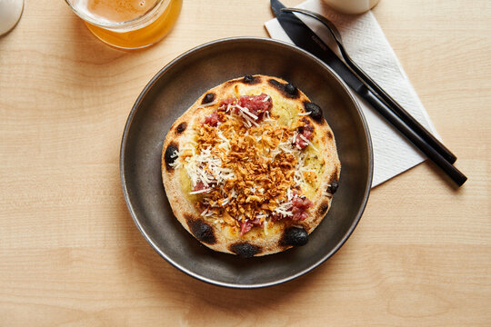 A mouthwatering flatbread adorned with a mixture of savory toppings sits on a rustic wooden table. Next to it, a glass of cold beverage and utensils are placed, completing the appetizing setup