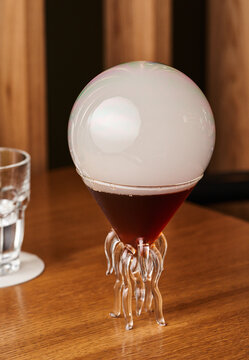 A striking cocktail topped with a large smoke bubble is presented in an artistic glass, with a faint glow and a hint of wood decor in the background during an evening out