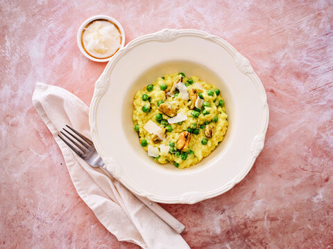 A delicious bowl of creamy risotto is filled with fresh green peas and mussels, garnished with cheese. A light cloth napkin and a small dish of seasoning accompany the meal