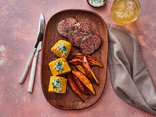 A beautifully arranged wooden platter holds slices of tender steak alongside golden corn and roasted sweet potato wedges. The setting evokes a warm and inviting dining atmosphere