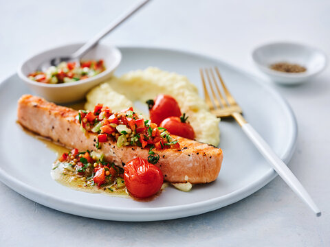 A beautifully plated salmon fillet topped with vibrant tomato salsa sits next to a serving of creamy mashed potatoes. The dish is garnished with fresh herbs, creating a mouthwatering presentation