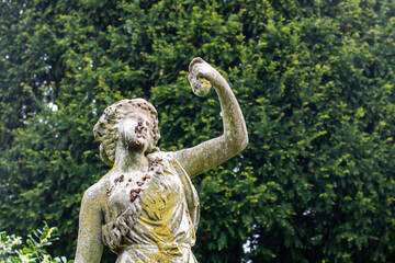 A statue of a woman with a cup in her hand. The statue is old and has a mossy appearance. The surrounding area is lush and green, with trees and bushes