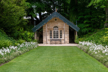 A small house with a green roof sits in a lush green field. The house is surrounded by a beautiful garden with white flowers. The garden is well-maintained and adds to the overall charm of the scene