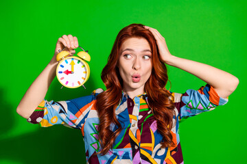 Cheerful young woman in vibrant printed shirt holding colorful alarm clock against bright green background