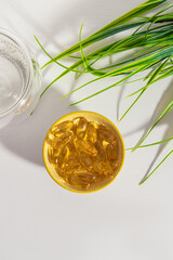 Transparent vitamin capsules in small bowl, glass of water and plant on white background. Vertical
