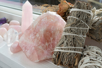 A close up image of a decorative white sage smudge stick adorned with dried roses and ribbon. Along side several polished rose quartz crystals and clear quartz tower.