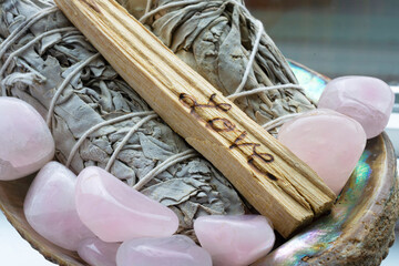 A close up image of two white sage smudge sticks surrounded polished rose quartz crystals in an abalone shell. This set up also included a smudge stick with the word love inscribed on it.