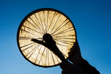 An image of a handheld homemade leather meditation drum with feather and drum stick being held against a bright blue sky.