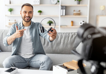 Cheery male vlogger recording video review of wireless headphones at home. Positive young blogger showing electronic device at camera, giving feedback, promoting new model of gadget
