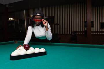 Young woman wearing stylish red sunglasses and a backwards baseball cap leans over a billiard...