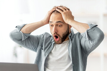 Terrified male freelancer looking at laptop screen, grabbing his head, making error in business project, missing deadline at home office. Young man having workplace stress, finding mistake in document