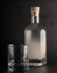 clear glass bottle, filled with white smoke and sealed with a wooden cork, stands beside an empty, clear glass tumbler. Both are set against a dark, textured black background.