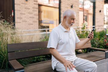 Fototapeta premium Thoughtful white bearded senior man with concerned expression holding smartphone, lost in thought as processes information on mobile phone screen, typing online message, searching, shopping online