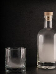 clear glass bottle, filled with white smoke and sealed with a wooden cork, stands beside an empty, clear glass tumbler. Both are set against a dark, textured black background.