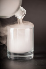 A close-up shot of a clear glass tumbler being filled with thick, white vapor. The vapor pours from a tilted glass bottle, overflowing the glass. The background is a soft-focus, dark gray.