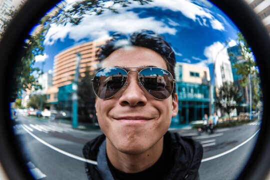 Man with Sunglasses Smiling Through a Fish-Eye Lens. A man wearing sunglasses smiles warmly through a fish-eye lens, capturing an urban backdrop with buildings and trees reflected in his glasses.