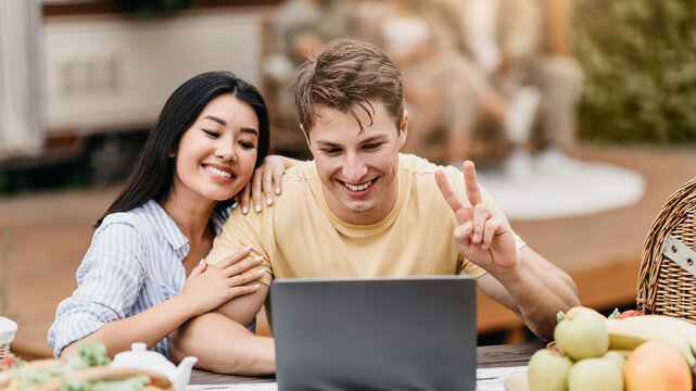 Millennial diverse couple using laptop computer near motorhome, video chatting online on summer camping trip. Happy Caucasian guy and his girlfriend communicating on pc during vacation