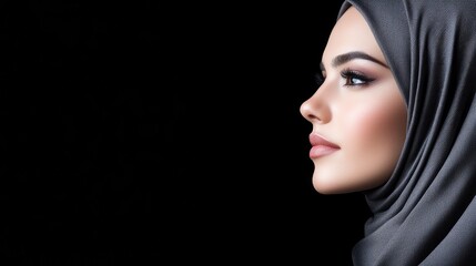 Fototapeta premium Young Muslim woman wearing a grey hijab poses in profile against a dark studio background.