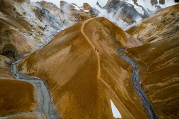 Scenic aerial drone view of a couple hikers in red jackets and foggy Kerlingarfjoll mountain path amidst desert and snowy terrain in Iceland