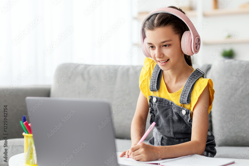 Poster studying at home. smiling pupil in pink headset sitting on couch at table using pc, writing essay or