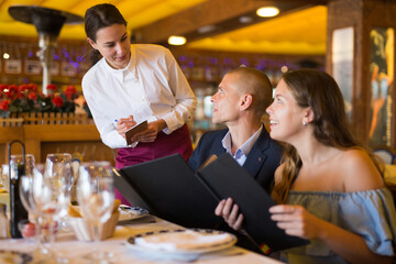 Attractive smiling woman waiter receiving order from guest in fashionable restaurant
