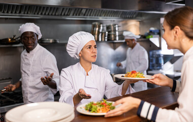 Portrait of smiling young woman chef working in restaurant kitchen, giving out ready salads s to waitress on order station