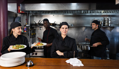 Portrait of focused woman head chef posing in modern restaurant kitchen, team of chefs working behind