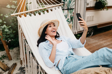 Obraz premium Cheerful young Asian woman lying in hammock near motorhome, having video chat on smartphone, waving at camera, enjoying summer vacation in countryside, copy space. Outdoor tourism concept