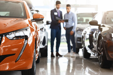 Customer middle eastern guy standing among brand new cars, having conversation with male sales assistant, looking at catalogue in dealer hands, checking auto features, full length, blurred background