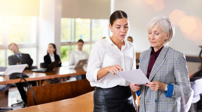 Elegantly dressed mature senior white female boss reading report file and discussing it with an active diligent young white female employee standing beside during business team meeting at office