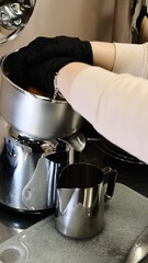 A barista in black disposable gloves squeezes fresh oranges on a professional juicer into a metal pitcher in a modern kitchen preparing a refreshing citrus beverage with vitamin C