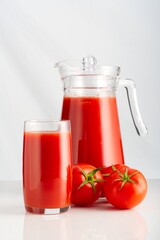 A glass of freshly squeezed tomato juice next to a stylish jug of vibrant red liquid. Ripe tomatoes surround them on a clean white background, showcasing their freshness.