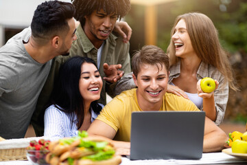 Group of diverse young friends using laptop while camping in countryside, communicating online, having fun. Multiracial millennial people spending time together outdoors on summer holidays