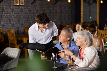 Elderly married couple relaxing in cozy restaurant. Waiter guy helps to sort out menu, choose original dish of non-traditional Oriental cuisine.