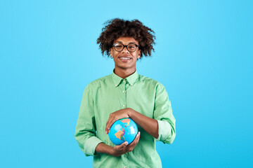 Happy african american teen guy protecting globe with his hands, posing on blue background and smiling at camera. Environmental protection, international tourism, tolerance for others concept