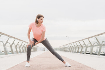 Young slim female jogger stretching warming-up her legs before running on the city bridge in...