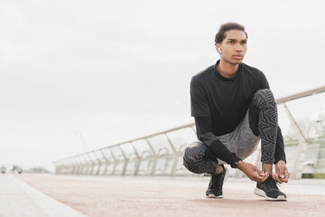 Young fit sporty african-american mixed-race male runner jogging on city bridge in sporty fitness outfit, tying his trainers before running workout outdoors.