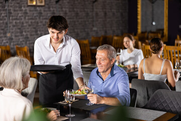 Young male waiter in uniform serving ordered dish to elderly couple in restaurant..