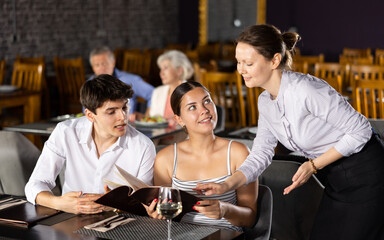 Friendly female owner of restaurant offering delicious dishes from menu to young couple sitting at table during romantic date