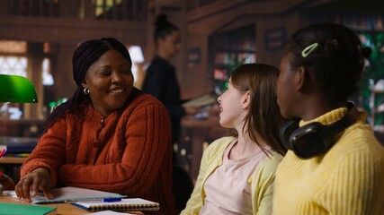 Obraz premium Diverse teen girls working with a tutor to complete class notes in a library, helps with learning and tutoring for the school subjects. Professor ensures diversity and child development. Camera B.