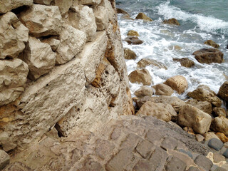 Naklejka premium Rustic coastal stone wall with textured rocks by the sea, waves crashing on weathered stones. Perfect for themes of nature, erosion, architecture, and coastal landscapes. 