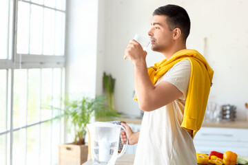 Young man with filter jug drinking fresh water in kitchen