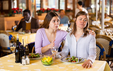 Caring woman calming her upset female friend while sitting together at table in cozy restaurant during dinner..