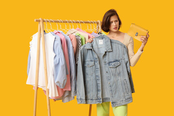 Thoughtful young woman near rack with different clothes choosing outfit on yellow background